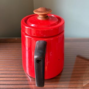 Pu&ograve; includere: Una teiera in ceramica rosso vivo con un manico nero e un pomello in legno sul coperchio. La teiera &egrave; cilindrica e poggia su una superficie di legno. &Egrave; un articolo per la cucina.