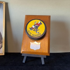 May include: A wooden plaque with a black and yellow hockey puck featuring a cartoon man sitting in a rocking chair. The puck says "Alte Herren" in black lettering. The plaque is on a small easel.