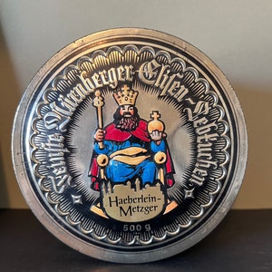May include: A silver round tin with a black and white image of a king sitting on a throne. The king is wearing a red and blue robe and a gold crown. The text around the image reads "Sankt-Künberger Elfen-Sektkuchen" and "Haeberlein-Metzger 500 g".