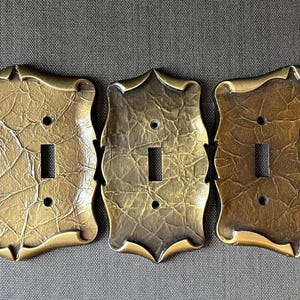 Pode incluir: Três tampas de interruptor de luz decorativas de latão com acabamento texturizado. As tampas têm um design decorativo e ornamentado ao redor das bordas. Uma tampa tem um único interruptor, outra tem um interruptor duplo e outra tem um interruptor duplo e uma tomada.