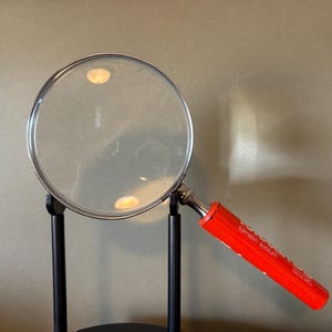 Puede incluir: Una lupa grande con una lente transparente y un mango rojo brillante. El mango tiene texto blanco que es difícil de leer. La lupa está montada en un soporte negro.