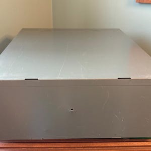 May include: A large, rectangular, grey metal storage box with a slightly textured surface. The box has a simple design with a few small, dark-coloured rectangular openings on the top. It sits on a wooden surface.