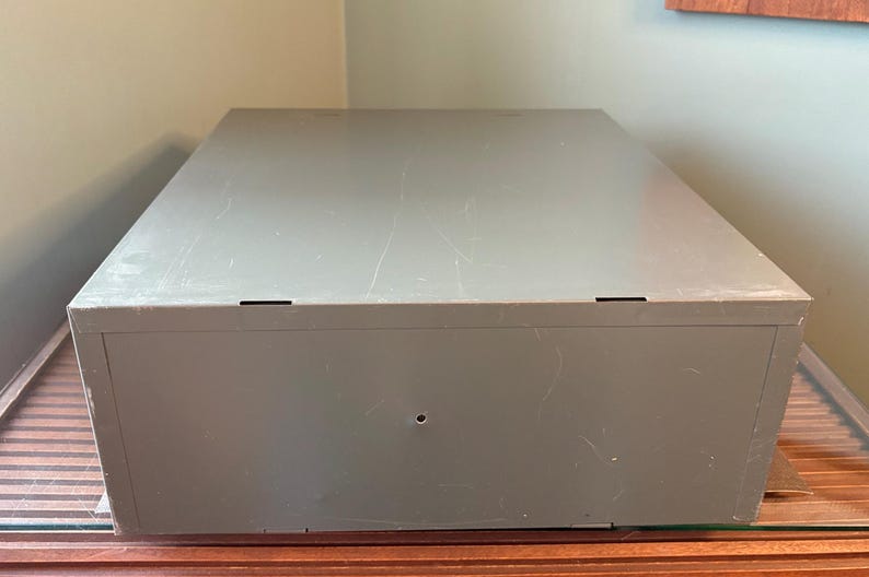May include: A large, rectangular, grey metal storage box with a slightly textured surface. The box has a simple design with a few small, dark-coloured rectangular openings on the top. It sits on a wooden surface.