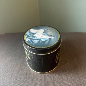 May include: A round, black tin with a gold rim and a lid featuring a colorful illustration. The tin's side has gold lettering that reads "Gifts". The lid shows a scene with people and furniture.