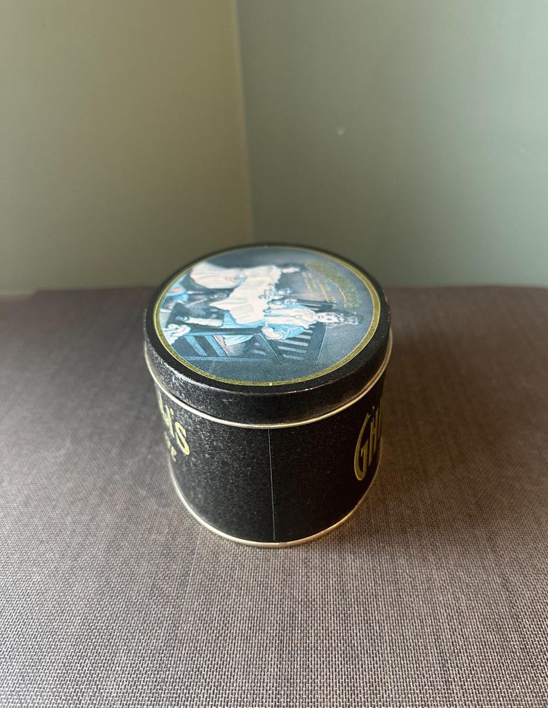 May include: A round, black tin with a gold rim and a lid featuring a colorful illustration. The tin's side has gold lettering that reads "Gifts". The lid shows a scene with people and furniture.