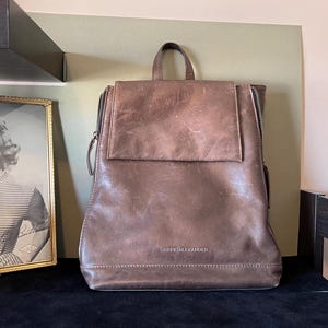 Könnte beinhalten: Ein brauner Lederrucksack mit Klappenverschluss und dem Schriftzug "Derek Alexander" auf der Vorderseite.