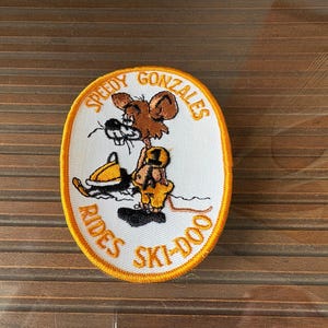 May include: Oval patch featuring Speedy Gonzales riding a yellow and black snowmobile. The patch has a white background with the text "Speedy Gonzales Rides Ski-Doo" in orange, outlined in yellow.