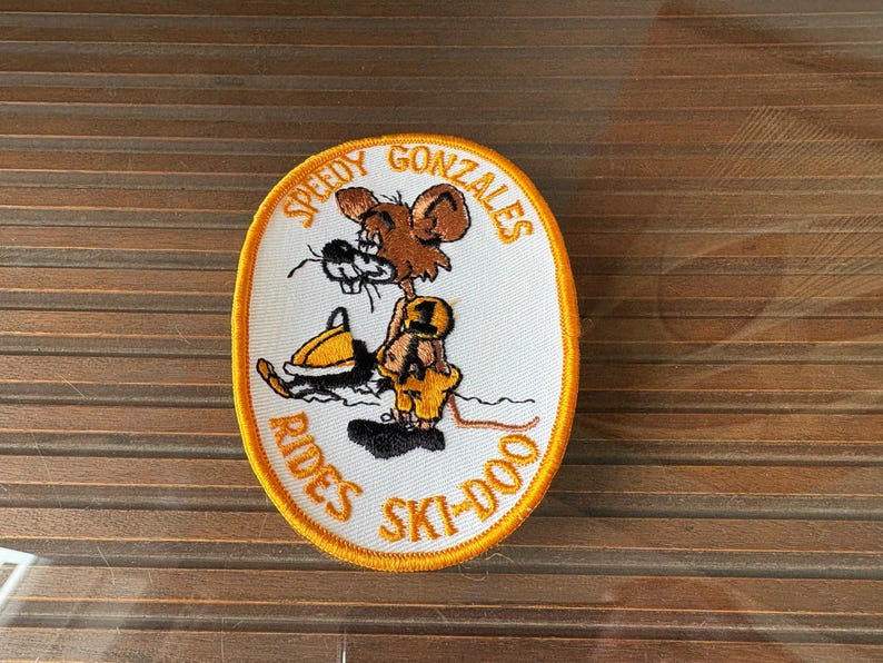 May include: Oval patch featuring Speedy Gonzales riding a yellow and black snowmobile. The patch has a white background with the text "Speedy Gonzales Rides Ski-Doo" in orange, outlined in yellow.