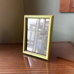Puede incluir: Un marco de fotos rectangular verde claro con un diseño estilo bambú. El marco está inclinado sobre una superficie de madera oscura, reflejando la luz. El interior del marco es negro, y el fondo es visible a través del cristal.