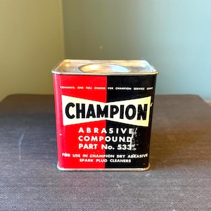 Puede incluir: Una lata de compuesto abrasivo Champion vintage. La lata rectangular presenta una combinación de colores rojo y negro con la palabra "CHAMPION" en letras blancas en negrita. El texto de la lata indica que es para usar en limpiadores de bujías abrasivos secos Champion.