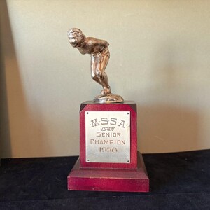 May include: A silver-toned trophy featuring a figure in a speed skating pose mounted on a dark red wooden base. A rectangular metal plaque on the front is engraved with the text MSSA OPEN SENIOR CHAMPION 1958 in black lettering.