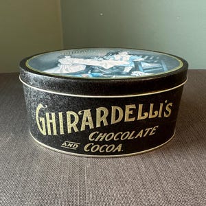 May include: Black Ghirardelli's Chocolate and Cocoa tin with a lid featuring a vintage illustration. The tin has gold lettering and a white rim. The tin is oval-shaped and sits on a brown surface.
