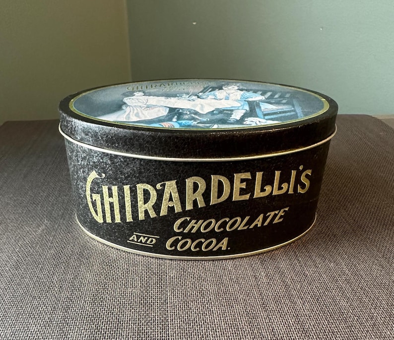 May include: Black Ghirardelli's Chocolate and Cocoa tin with a lid featuring a vintage illustration. The tin has gold lettering and a white rim. The tin is oval-shaped and sits on a brown surface.