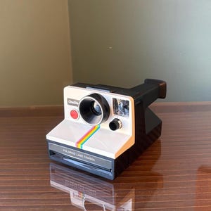 Peut inclure: Un appareil photo Polaroid Land Camera vintage, blanc avec un dessus et une base noirs. Une bande arc-en-ciel orne le devant, avec les mots "Polaroid Land Camera" imprimés en dessous. L'appareil photo a un bouton rouge et un objectif noir.