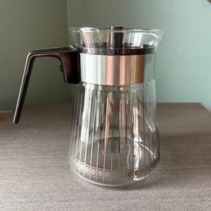 Może przedstawiać: Szklany dzbanek do kawy z czarnym uchwytem i srebrną obręczą wokół górnej części. Dzbanek ma fakturowany wzór i srebrną obręcz wokół górnej części.