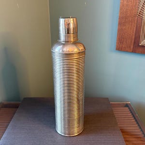 Può includere: Un'alta shaker per cocktail argentata con un design a coste. Lo shaker ha un tappo e un corpo cilindrico. È fatto di metallo e poggia su una superficie scura. Lo sfondo è verde e blu tenui.