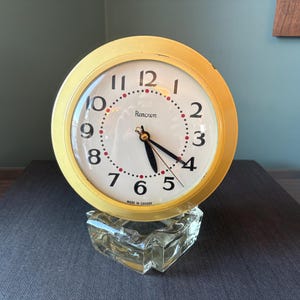 Peut inclure: Une horloge jaune vintage avec un cadran blanc, des chiffres noirs et des points rouges indiquant les minutes. L'horloge, marquée "Renown", repose sur une base en verre transparente et géométrique. Les aiguilles indiquent environ 4h27.