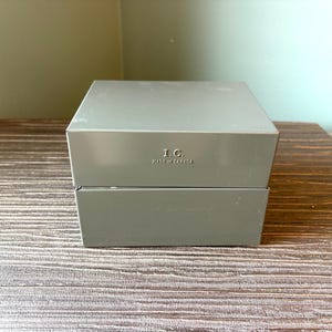以下が含まれることがあります： 蓋付きの正方形のマットグレーの収納ボックス。蓋の前面には「I.C.」というエンボス加工の文字があり、その下に「MADE IN CANADA」という言葉が中央揃えで配置されています。ボックスは滑らかでしっかりとした構造で、全体的に均一な外観です。