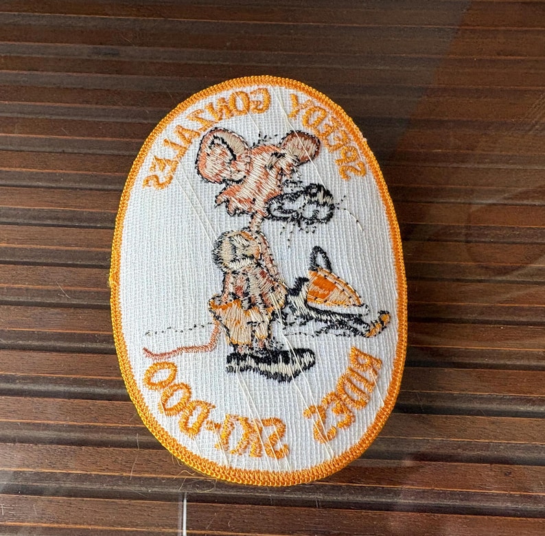 May include: Oval-shaped embroidered patch with a cartoon mouse riding a motorcycle. The patch features orange and white colors, with text around the border. The mouse is wearing a helmet and the motorcycle is orange. The text reads "Speedy Gonzales" and "Ride or Die".