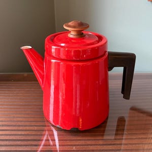 May include: A red enamel teapot with a wooden knob on the lid and a dark brown handle. The spout has a silver tip. The teapot is sitting on a wooden surface.