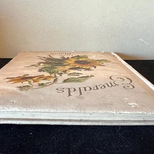 May include: A white book with a floral design on the cover. The book is titled "Emeralds" and has a white spine.