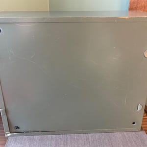 May include: An olive-green metal cabinet, rectangular in shape, showing signs of wear. The cabinet has a plain design with a flat top and sides, and includes small circular and D-shaped hardware. The surface is marked with scratches.