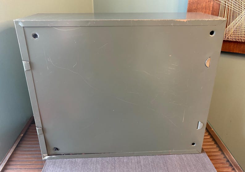 May include: An olive-green metal cabinet, rectangular in shape, showing signs of wear. The cabinet has a plain design with a flat top and sides, and includes small circular and D-shaped hardware. The surface is marked with scratches.