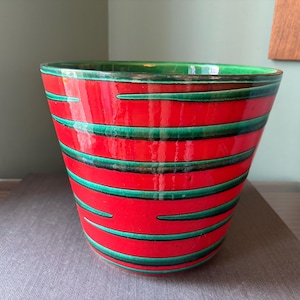 May include: A vibrant red ceramic planter with horizontal green stripes. The planter has a glossy finish and a green rim. The design is a bold, eye-catching statement piece for indoor or outdoor use. The planter is approximately 12 inches tall.