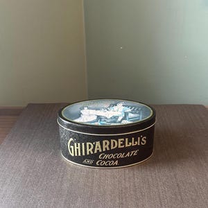 May include: A round Ghirardelli's Chocolate and Cocoa tin. The tin is black with gold lettering and a detailed illustration on the lid. The lid features a scene with people. The tin is sitting on a brown surface.