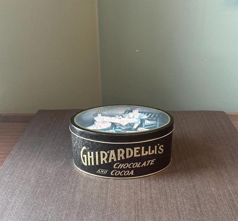 May include: A round Ghirardelli's Chocolate and Cocoa tin. The tin is black with gold lettering and a detailed illustration on the lid. The lid features a scene with people. The tin is sitting on a brown surface.