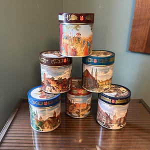 May include: Seven vintage cylindrical tins with lids are stacked. The tins display colourful illustrations of European town scenes and figures. The lids and rims are in various colours, including red, blue, and brown.