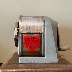 Puede incluir: Un sacapuntas vintage plateado con una etiqueta roja y blanca que dice "Deluxe Giant Automatic Pencil Sharpener Co. Toronto, Canada" y un logotipo rojo que dice "Apco".