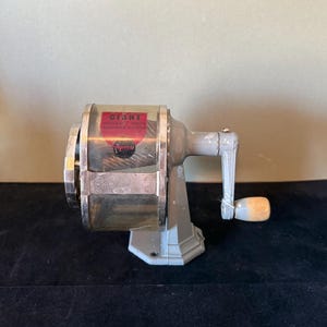 Op de afbeelding: Een vintage grijze puntenslijper met een zilveren en rood label met de tekst "GIANT". De puntenslijper heeft een slinger met een houten knop en een zilveren mesbehuizing. De basis is lichtgrijs.