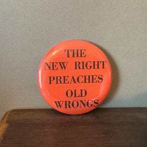 May include: A circular, orange badge with the black text "THE NEW RIGHT PREACHES OLD WRONGS". The badge is displayed on a wooden surface, set against a pale green backdrop.
