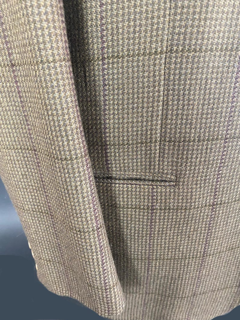 May include: Close-up of a tweed blazer in shades of brown and gray with a subtle purple plaid pattern. The jacket features a notched lapel and a pocket, showcasing the tailored design and textured fabric.