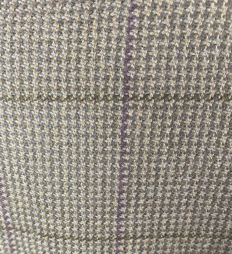 May include: Close-up of a textured fabric with a houndstooth pattern in shades of beige, gray, and a thin purple stripe. The weave is tight, creating a detailed and visually interesting surface. The fabric appears to be a woven material, possibly wool or a wool blend.