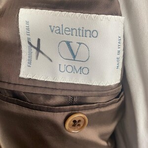May include: Close-up of a brown Valentino Uomo suit jacket interior. The label reads "valentino UOMO" with a "V" logo. The jacket has a brown satin lining and a button closure.