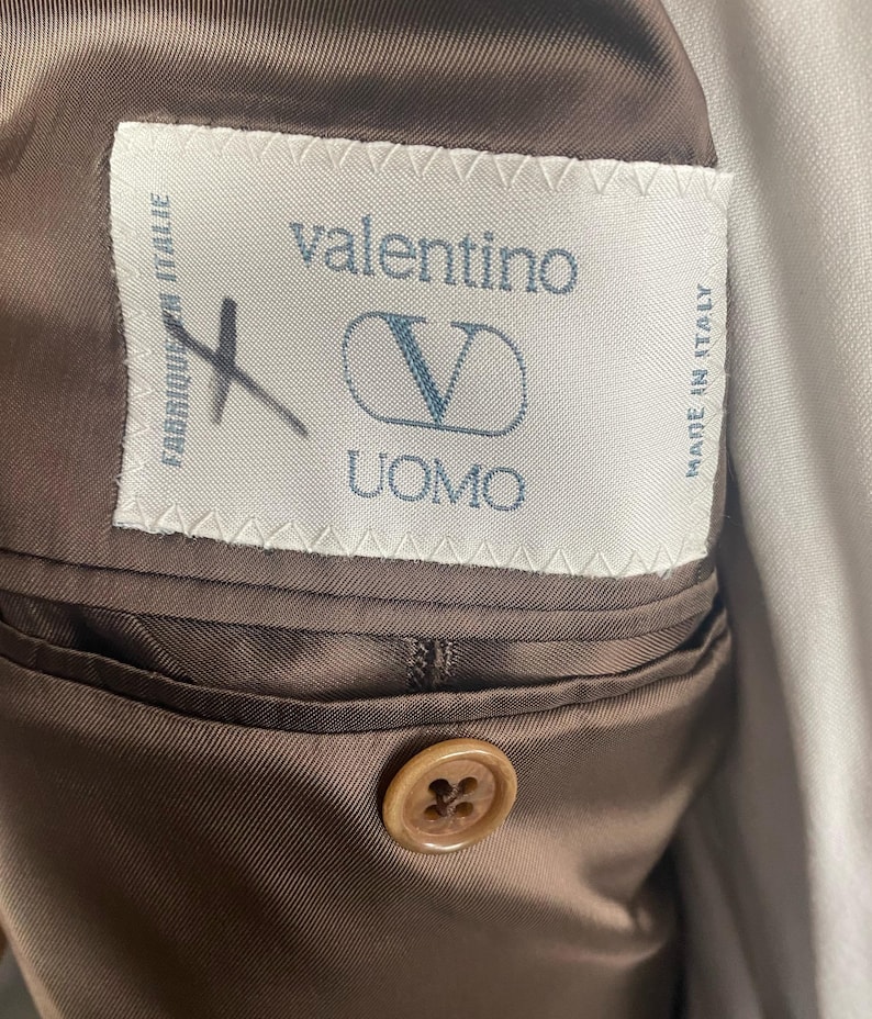 May include: Close-up of a brown Valentino Uomo suit jacket interior. The label reads "valentino UOMO" with a "V" logo. The jacket has a brown satin lining and a button closure.