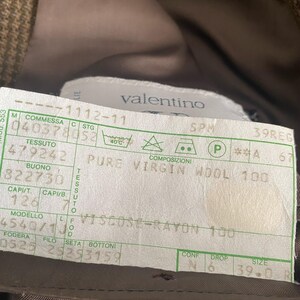 May include: A close-up of a Valentino label, featuring text indicating the garment's composition: Pure Virgin Wool 100 and Viscose-Rayon 100. The label also includes size information and care instructions. The background is a brown fabric.