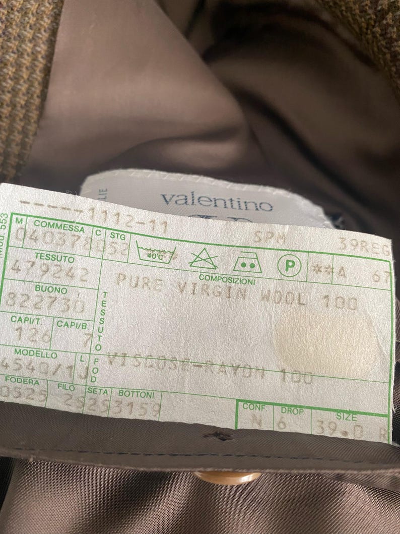 May include: A close-up of a Valentino label, featuring text indicating the garment's composition: Pure Virgin Wool 100 and Viscose-Rayon 100. The label also includes size information and care instructions. The background is a brown fabric.