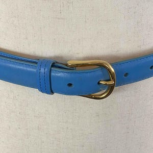 May include: A light blue leather belt with a gold-tone buckle. The belt is a classic style, with a simple design and a polished finish. The belt has a width of approximately 1 inch.