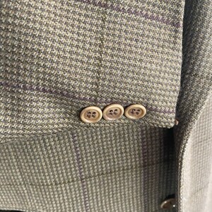 May include: Close-up of a tweed blazer in shades of green and brown with a subtle purple plaid pattern. The sleeve features three round, light-colored buttons. The fabric has a textured appearance.