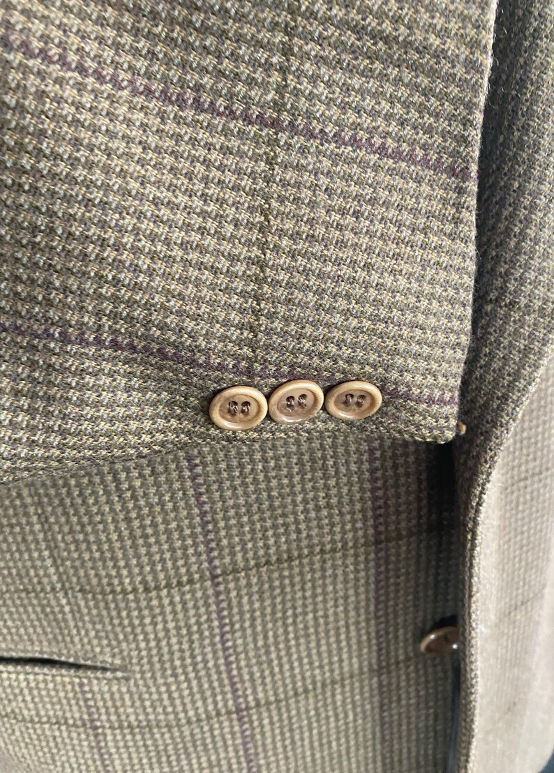 May include: Close-up of a tweed blazer in shades of green and brown with a subtle purple plaid pattern. The sleeve features three round, light-colored buttons. The fabric has a textured appearance.