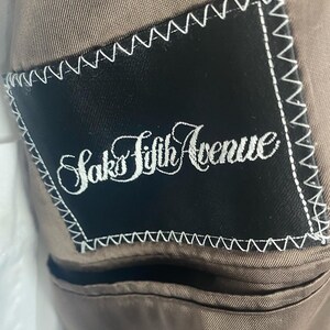 May include: Close-up of a brown suit jacket interior. A black label with white stitching is sewn into the lining. The label reads "Saks Fifth Avenue" in elegant script.