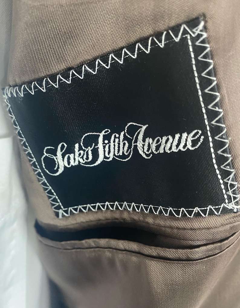 May include: Close-up of a brown suit jacket interior. A black label with white stitching is sewn into the lining. The label reads "Saks Fifth Avenue" in elegant script.