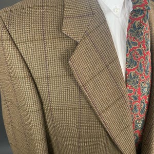 May include: A brown tweed blazer with a subtle plaid pattern. The jacket is paired with a white dress shirt and a red paisley tie. The blazer's texture and color suggest a classic, tailored style.