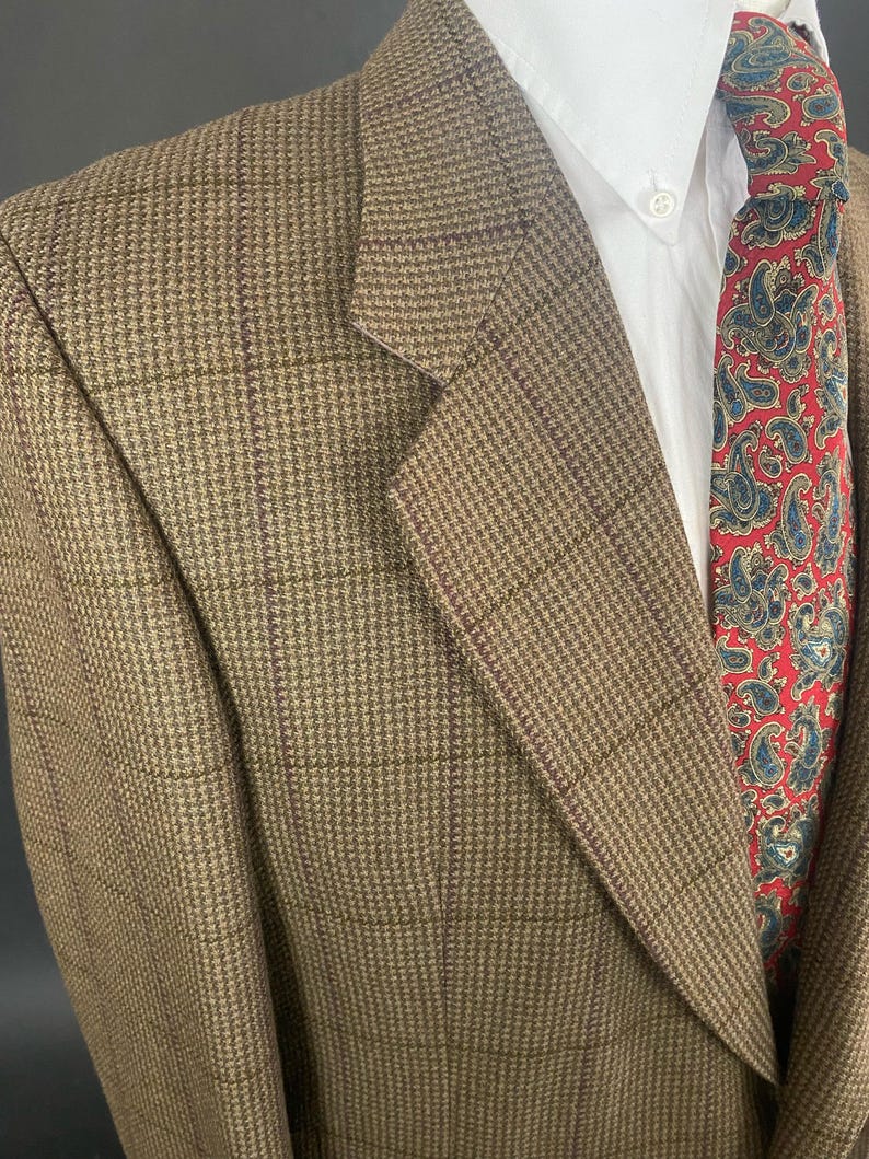 May include: A brown tweed blazer with a subtle plaid pattern. The jacket is paired with a white dress shirt and a red paisley tie. The blazer's texture and color suggest a classic, tailored style.