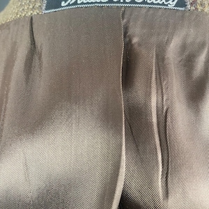May include: Close-up of the inside of a brown tweed jacket. The lining is a smooth, satin-like material. A black tag with the words "Made in Italy" is sewn into the collar.