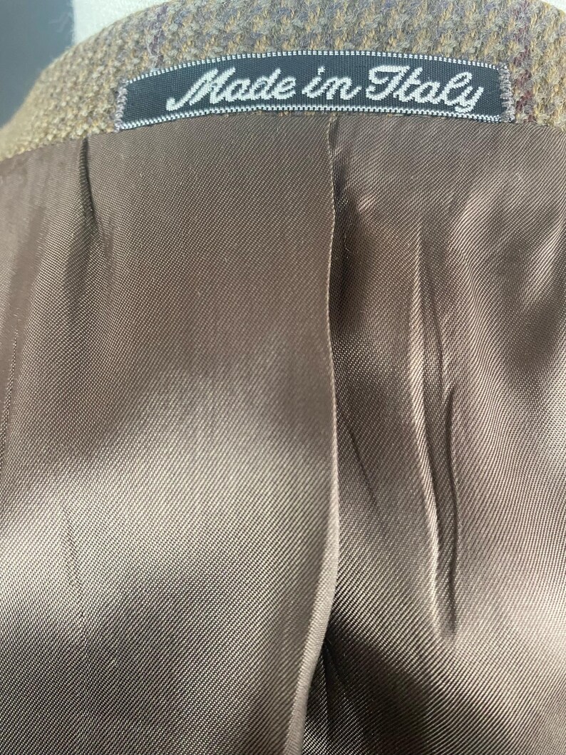 May include: Close-up of the inside of a brown tweed jacket. The lining is a smooth, satin-like material. A black tag with the words "Made in Italy" is sewn into the collar.