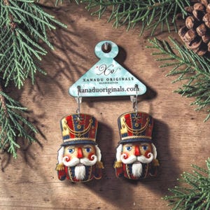 May include: A pair of nutcracker earrings featuring red, blue, and gold uniforms with white beards, attached to a light blue display card labeled Xanadu Originals and xanaduoriginals.com. The earrings hang from silver-toned metal hooks.